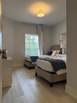 A bedroom with two beds, a window with a view of greenery, a ceiling light, and a bedside table with a lamp. - Lakeside Serenity at Worldquest (Orlando)