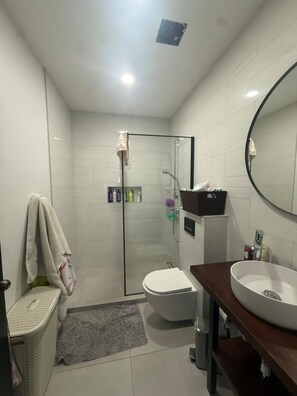 A modern bathroom with a glass shower enclosure, a toilet, a sink with a mirror, and a laundry basket. - Wayaca 3br villa with private pool (Oranjestad)