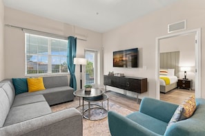 A modern hotel room with a sofa, armchair, a small round table, a cabinet, a bed, and a TV on the wall. - Work and Play, Spacious 3BR at Vista Cay (Orlando)