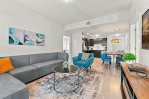 A modern living room with a grey sofa, blue armchairs, and a coffee table. There is a kitchen in the background with dark cabinets and a dining area with blue chairs. - Comfortable 3BR Family Getaway, Top Location (Orlando)