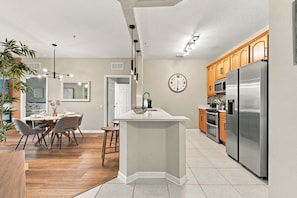 A modern kitchen with a central island, wooden cabinets, stainless steel appliances, and a dining area with a round table and chairs. - Vista Cay 3br, Comfort & Prime Location, Sleeps 7 (Orlando)