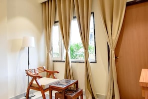 A room with a wooden chair, a small table, a floor lamp, and a window with curtains. - The Nest (Varanasi)