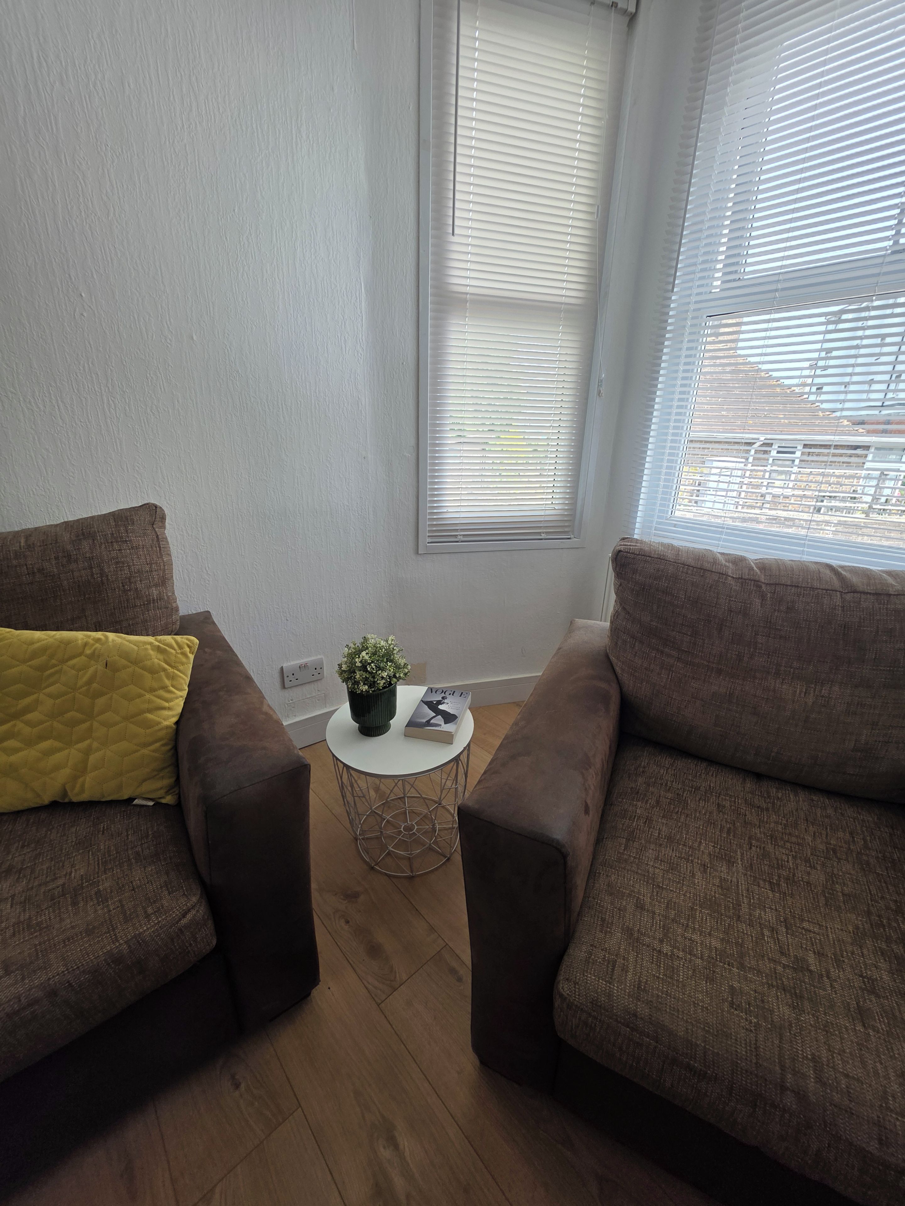 A room with two brown sofas, a small round table with a plant and a magazine, and a window with blinds.