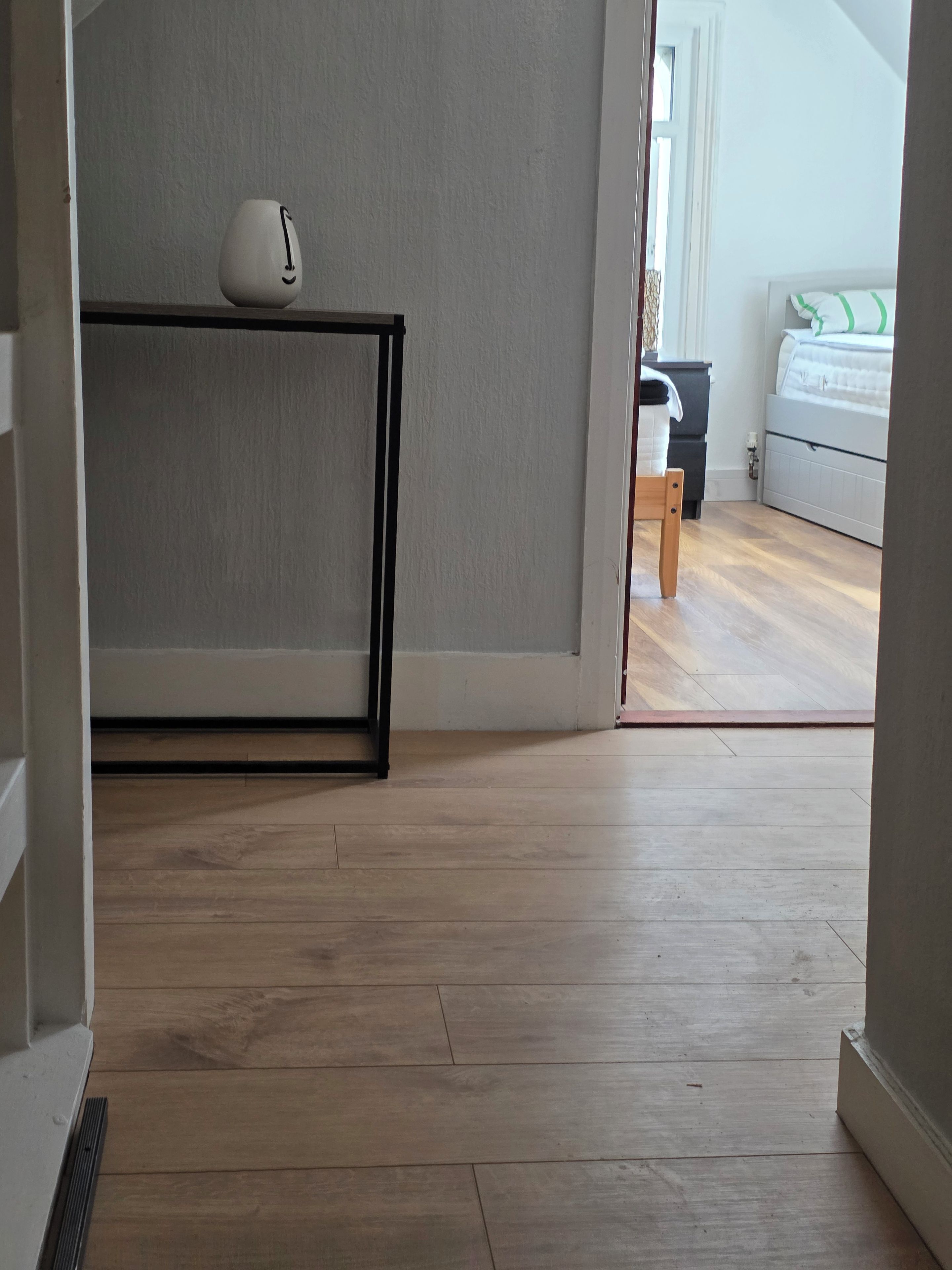 Corredor com piso de madeira, parede branca e porta que dá para um quarto com cama e cômoda.