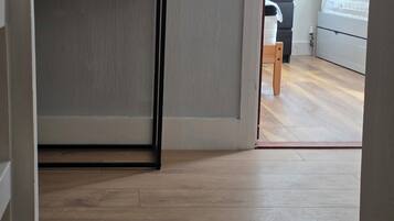 Corredor com piso de madeira, parede branca e porta que dá para um quarto com cama e cômoda.