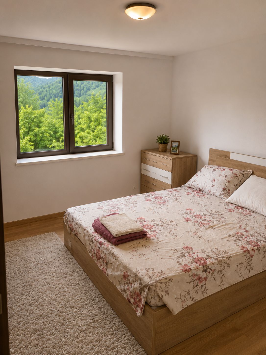 A bedroom with a bed, a nightstand, a dresser, a window with a view of a forest, and a ceiling light.