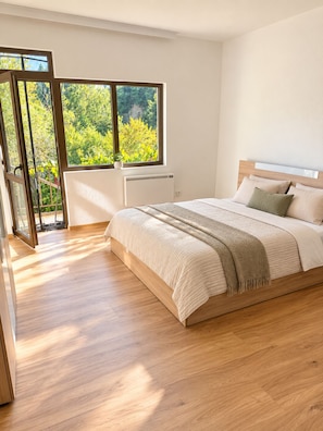 A bedroom with a large bed, a window with a view of greenery, and a sliding door leading to an outdoor area.