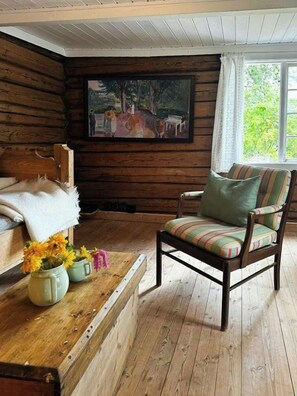 A cabin interior with a wooden floor, a bed with a white blanket, a chair with a striped cushion, a painting on the wall, and a window with a view of greenery. - Idyll, history and nature – all in one place (Hornnes)