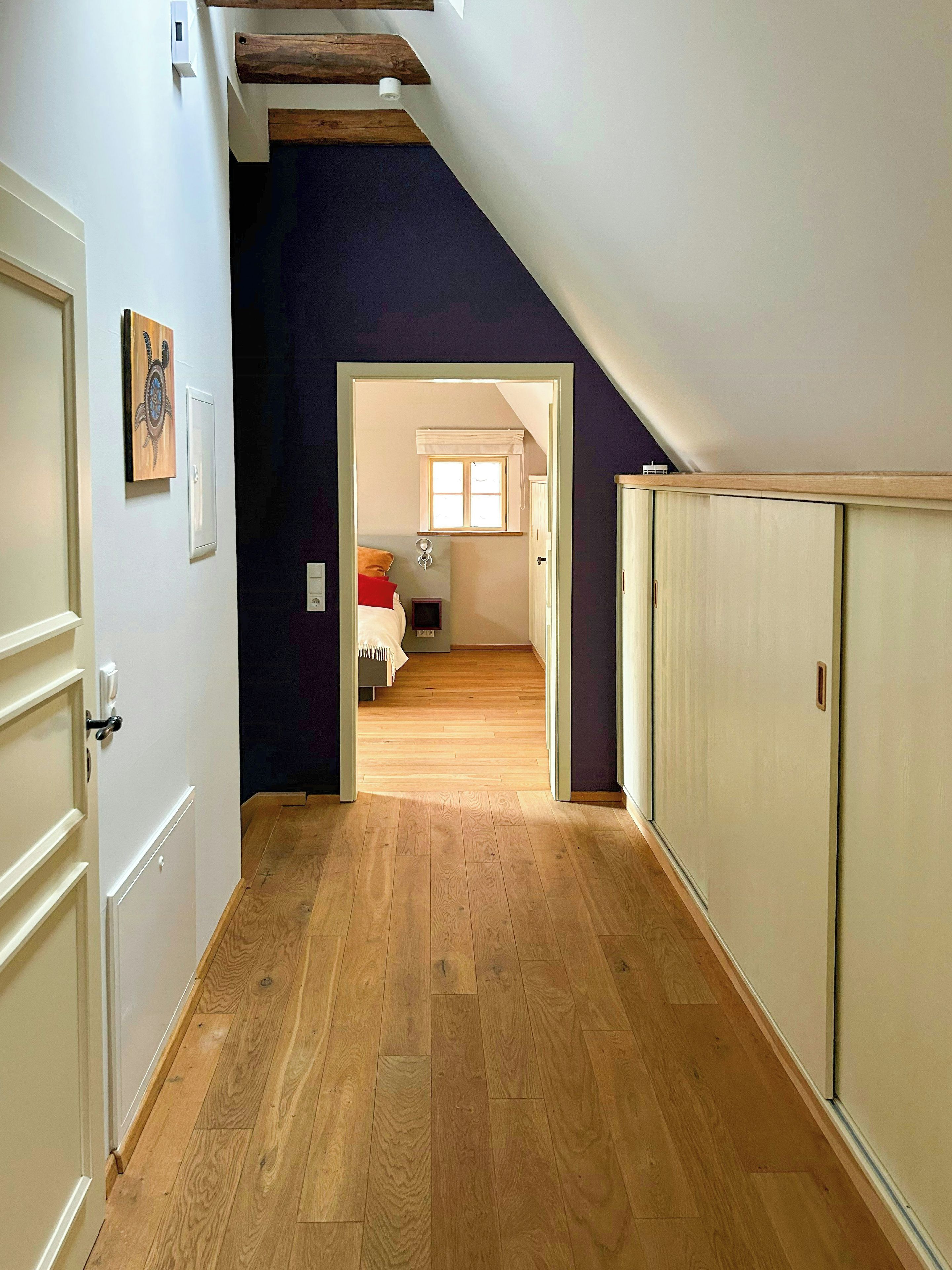 A hallway with wooden flooring leading to a bedroom with a window and a bed.