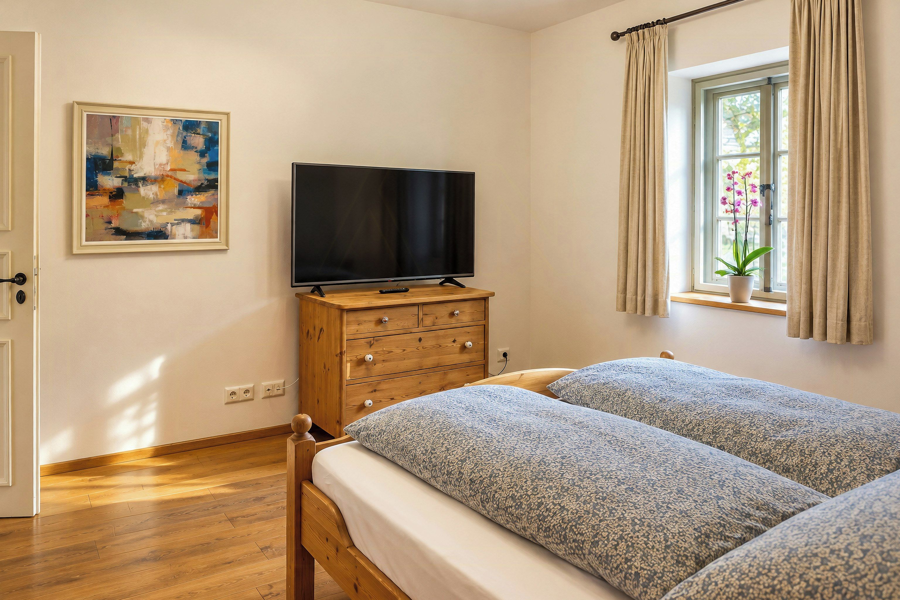 Een slaapkamer met een bed, een televisie op een houten dressoir, een schilderij aan de muur en een raam met gordijnen.