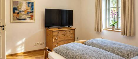 A bedroom with a bed, a television on a wooden dresser, a painting on the wall, and a window with curtains.