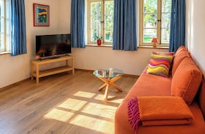 Un soggiorno con una TV a schermo piatto su un supporto in legno, un tavolino da caffè in vetro, un divano arancione con cuscini colorati e ampie finestre con tende blu.