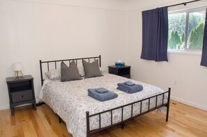 A bedroom with a metal bed frame, floral bedding, two pillows, a bedside lamp, a nightstand, a window with curtains, and a view of trees. - Farm stay in the heart of Metchosin, near Victoria and outdoor adventures (Victoria)