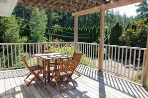A wooden deck with a table and chairs, surrounded by a white railing, overlooking a forested area.