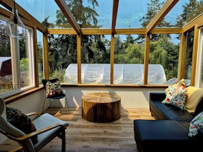 A conservatory with a wooden floor, a round wooden table, a sofa with floral cushions, and a chair with a floral cushion. - Farm stay in the heart of Metchosin, near Victoria and outdoor adventures (Victoria)