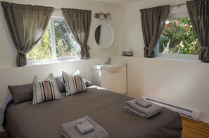 A bedroom with a bed, pillows, a bedside table, a mirror, a window with curtains, and a view of greenery outside.