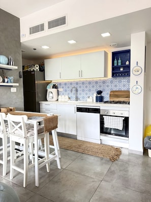 A modern kitchen with white cabinetry, a built-in oven, and a dining area with white chairs. - Little greece - Charming Haifa Downtown Apartment (Haifa)