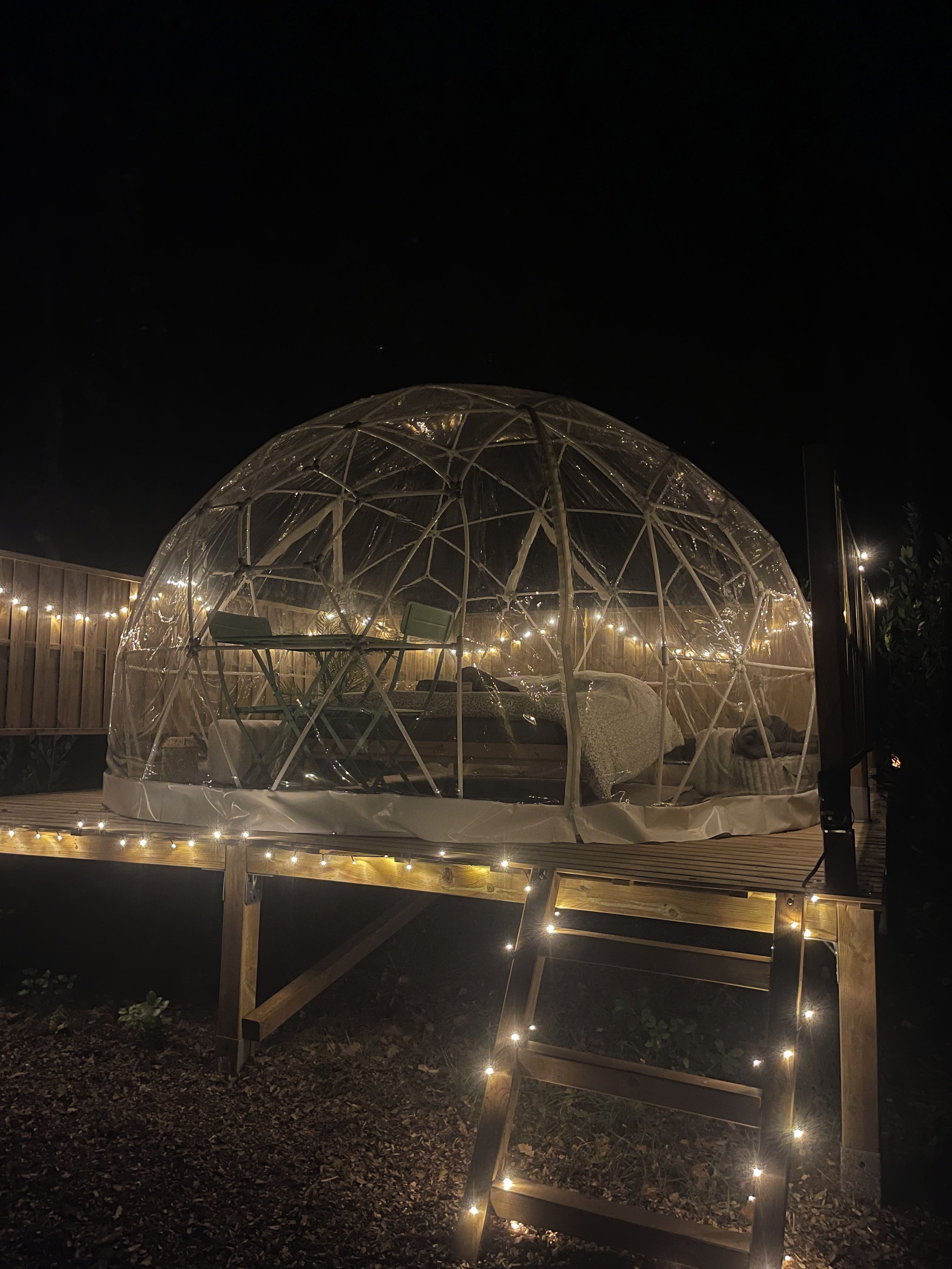 Una estructura de domo geodésico con techo transparente, rodeada por una terraza de madera e iluminada por luces colgantes.