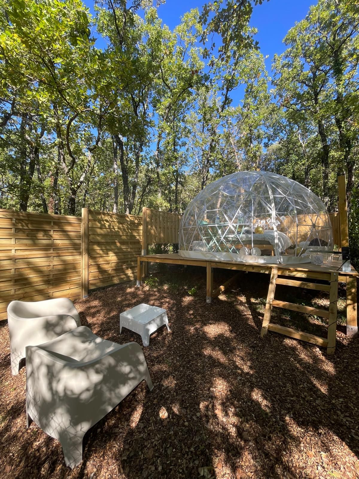 Une structure en forme de dôme géodésique transparente, avec une table et des chaises à l’extérieur, entourée d’arbres.
