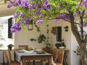 An outdoor dining area with a table set for two, surrounded by chairs, under a tree with purple flowers. - Rural cortijo with pool and Mediterranean views (Canillas de Aceituno)