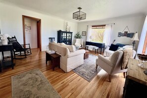 A living room with a wooden floor, a sofa, a chair, a cabinet, and a window with curtains. - Two-story farmhouse on a working alfalfa farm, minutes from the wine district (Walla Walla)