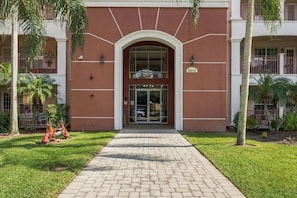 A building entrance with a brick pathway leading to a glass door, palm trees, and a fire hydrant. - Upscale Vista Cay 3BR With Resort Pool View (Orlando)