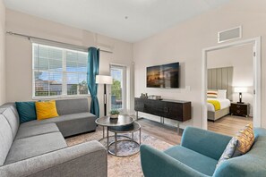 A modern hotel room with a sofa, armchair, a small round table, a cabinet, a bed, and a TV mounted on the wall. - Work and Play Spacious 3BR at Vista Cay (Orlando)