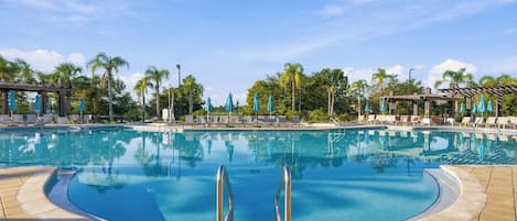 A large outdoor swimming pool with a clear sky, surrounded by palm trees and lounge chairs.