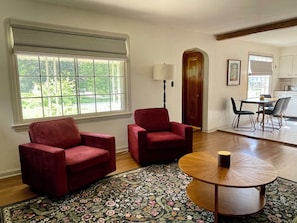 A living room with two red armchairs, a coffee table, a patterned rug, a window with a view of greenery, and a door leading to a kitchen. - Charming Vintage renovated 2 story Cape Cod home near Boardman Shopping (Youngstown)