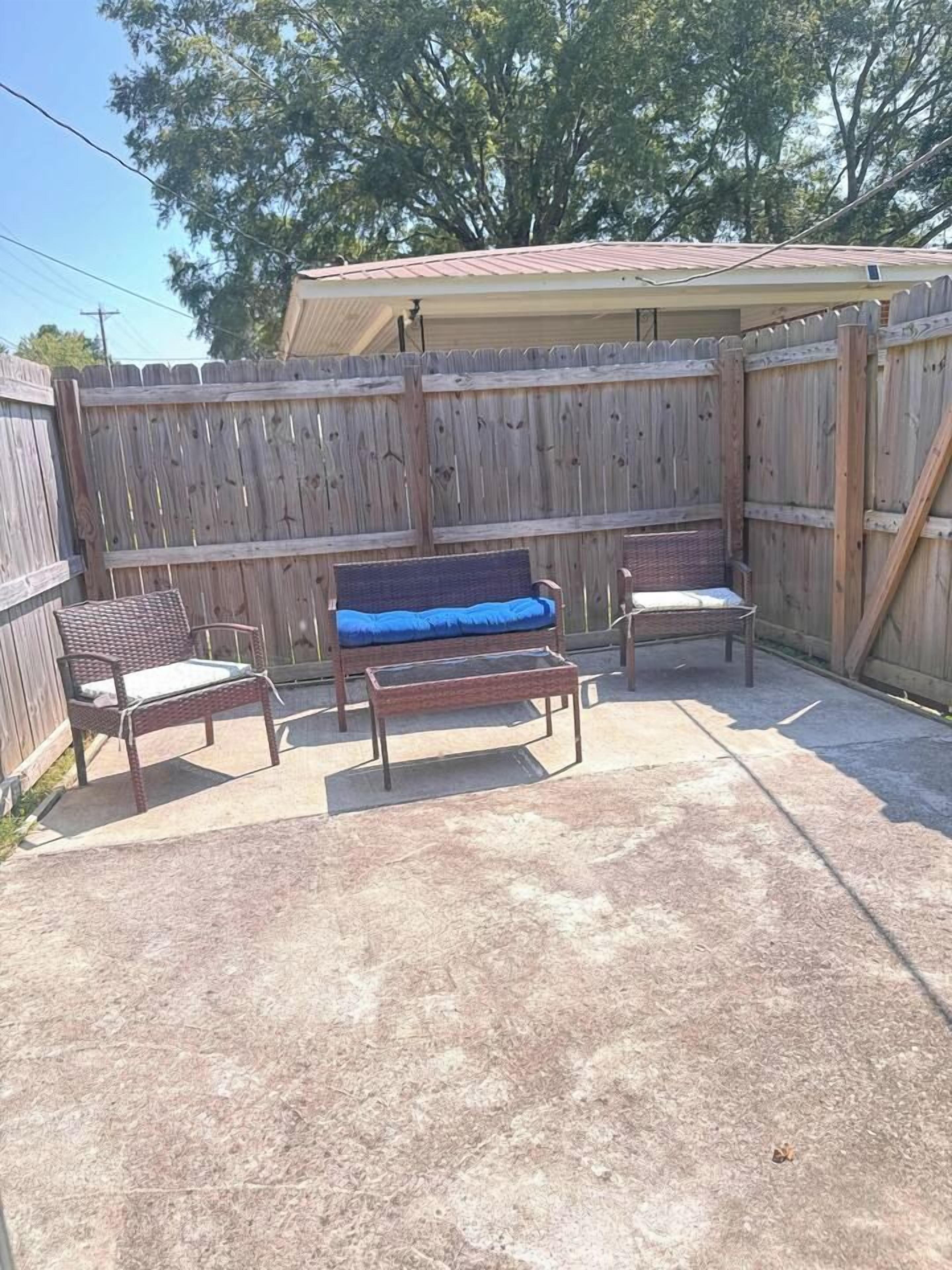 A patio area with a wooden fence, a covered structure, and outdoor furniture including a bench and two chairs.