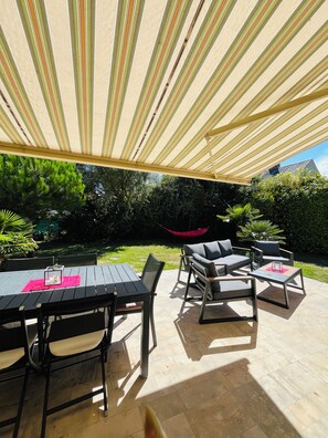 Un patio exterior con mesa y sillas, una hamaca y una zona con sombra gracias a un dosel a rayas.