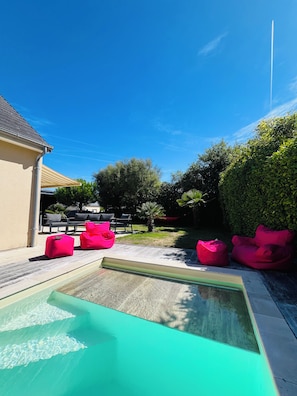 Ein Schwimmbecken mit klarem, blauen Wasser, umgeben von Sitzgelegenheiten im Freien mit leuchtend pinkfarbenen Sitzsäcken, sowie ein gepflegter Garten mit verschiedenen Pflanzen und einer Hängematte.