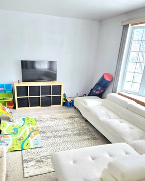 A living room with a white sofa, a television on a wooden stand, a colorful play mat, and a yoga mat leaning against the wall.