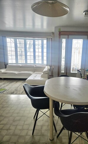 A room with a white sofa, a round table with chairs, and large windows with a view of trees.