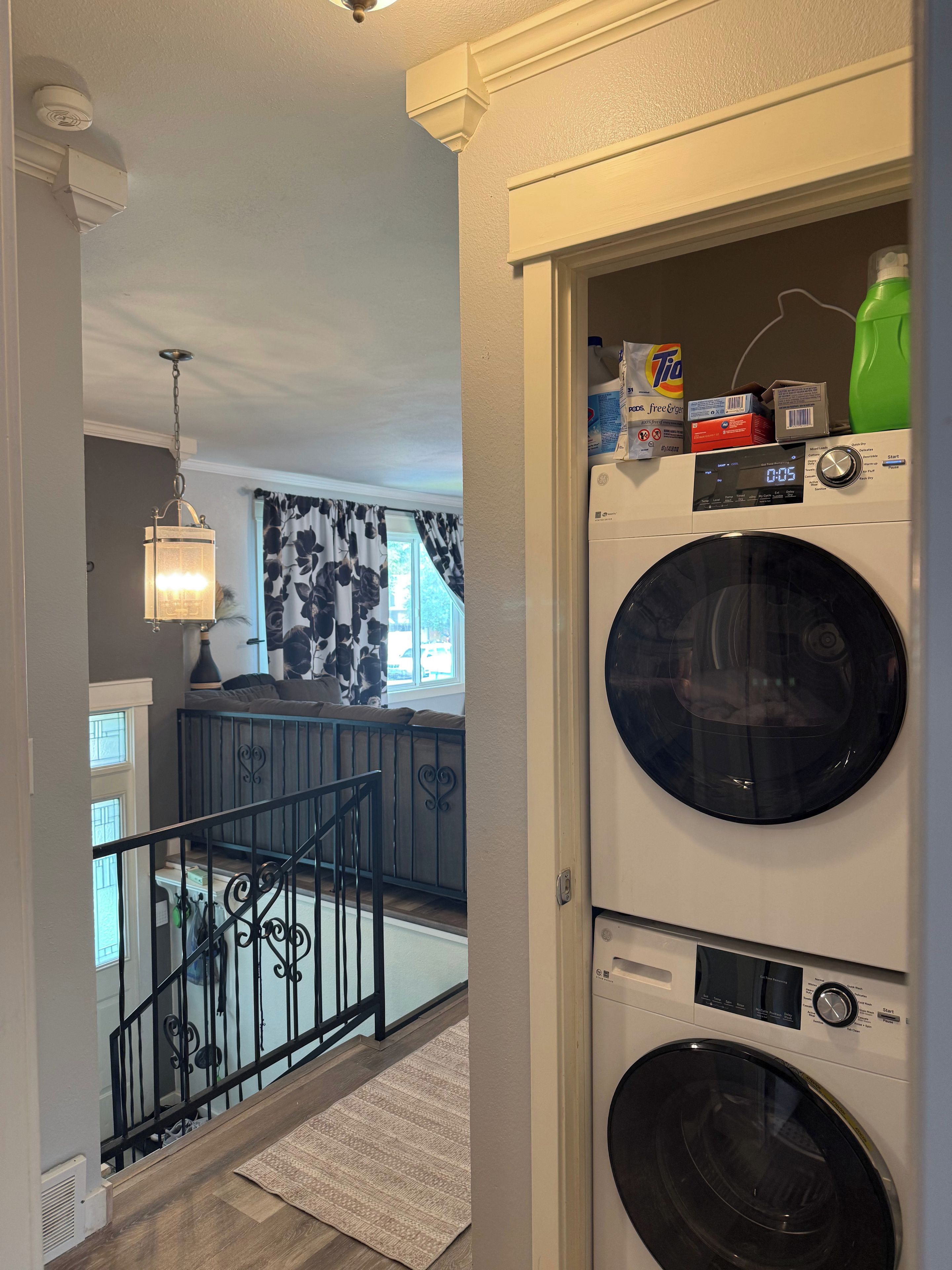 A laundry room with stacked washers and dryers, a hanging laundry lamp, and a window with curtains.