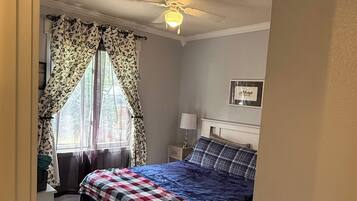A bedroom with a bed, a ceiling fan, a window with curtains, a bedside table with a lamp, and a framed picture on the wall.