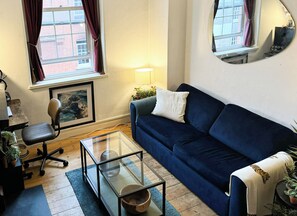 A living room with a dark blue sofa, a glass coffee table, a framed picture on the wall, a lamp, and a window with curtains. - Artist's 1 Bed Apartment in Westminster / Central London (London)