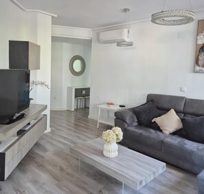 Ein modernes Wohnzimmer mit Flachbildfernseher, grauem Sofa, weißem Couchtisch und einer Vase mit Blumen.