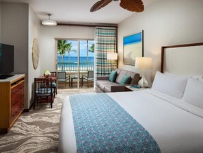 Ein Hotelzimmer mit einem großen Bett, einem Fernseher, einem Schreibtisch mit Stühlen und Blick auf den Strand.