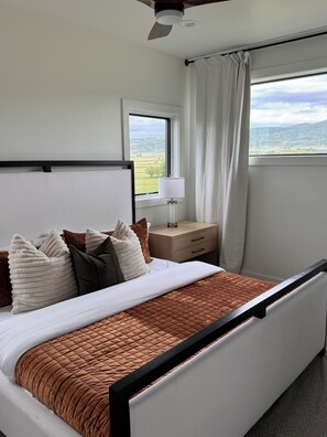 Un dormitorio moderno con cama, mesita de noche, lámpara y vistas al campo a través de una ventana.