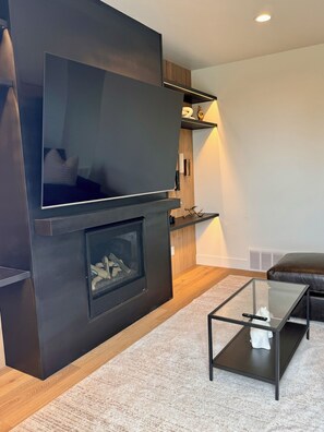 Una sala de estar moderna con un televisor de pantalla plana montado en una pared oscura, una mesa de centro de vidrio, un sofá de cuero marrón y una chimenea con leña.