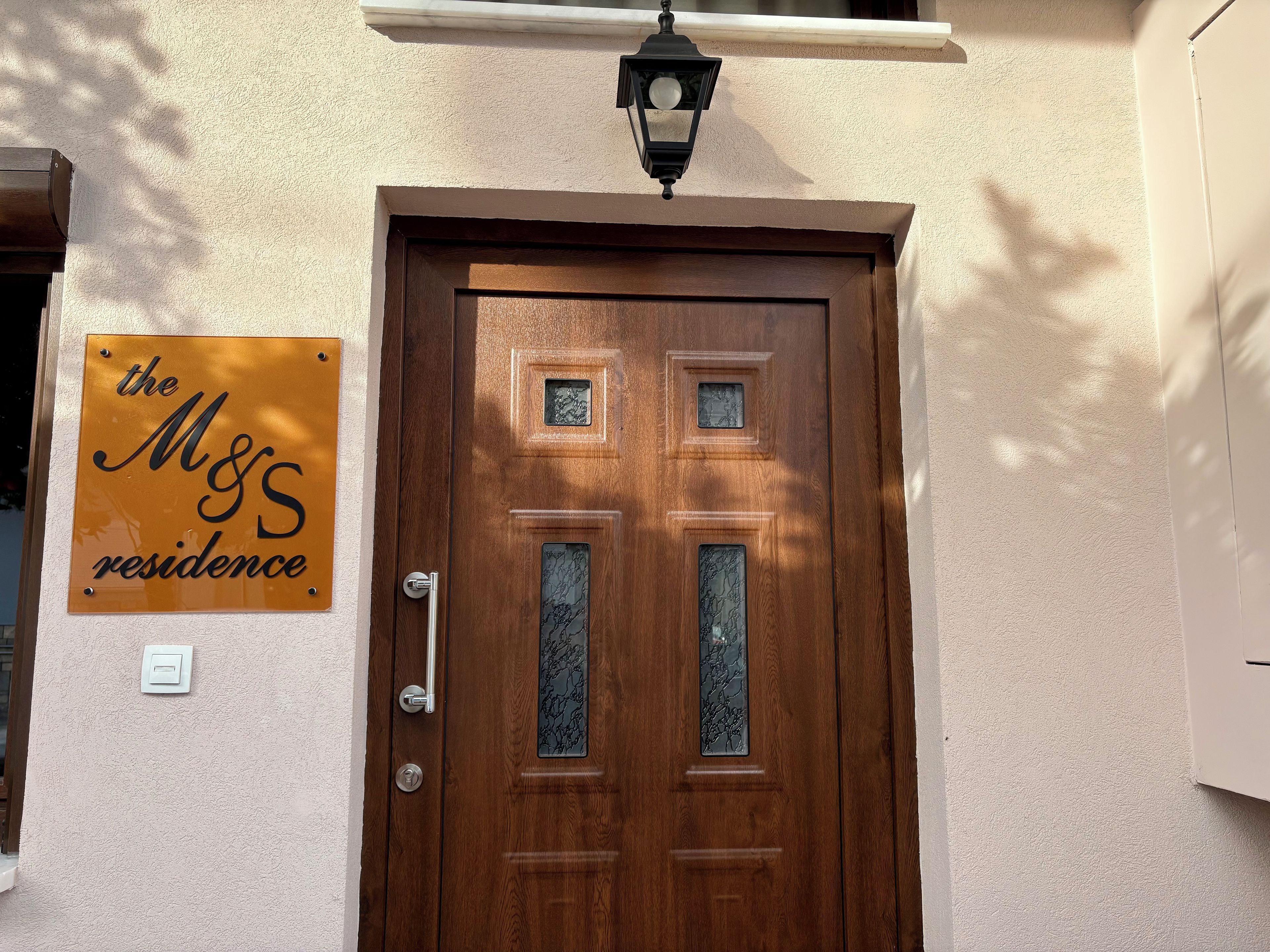 A wooden door with a metal handle and a glass panel, a wall-mounted light fixture above, and a sign reading 'the M&S residence' on the wall.