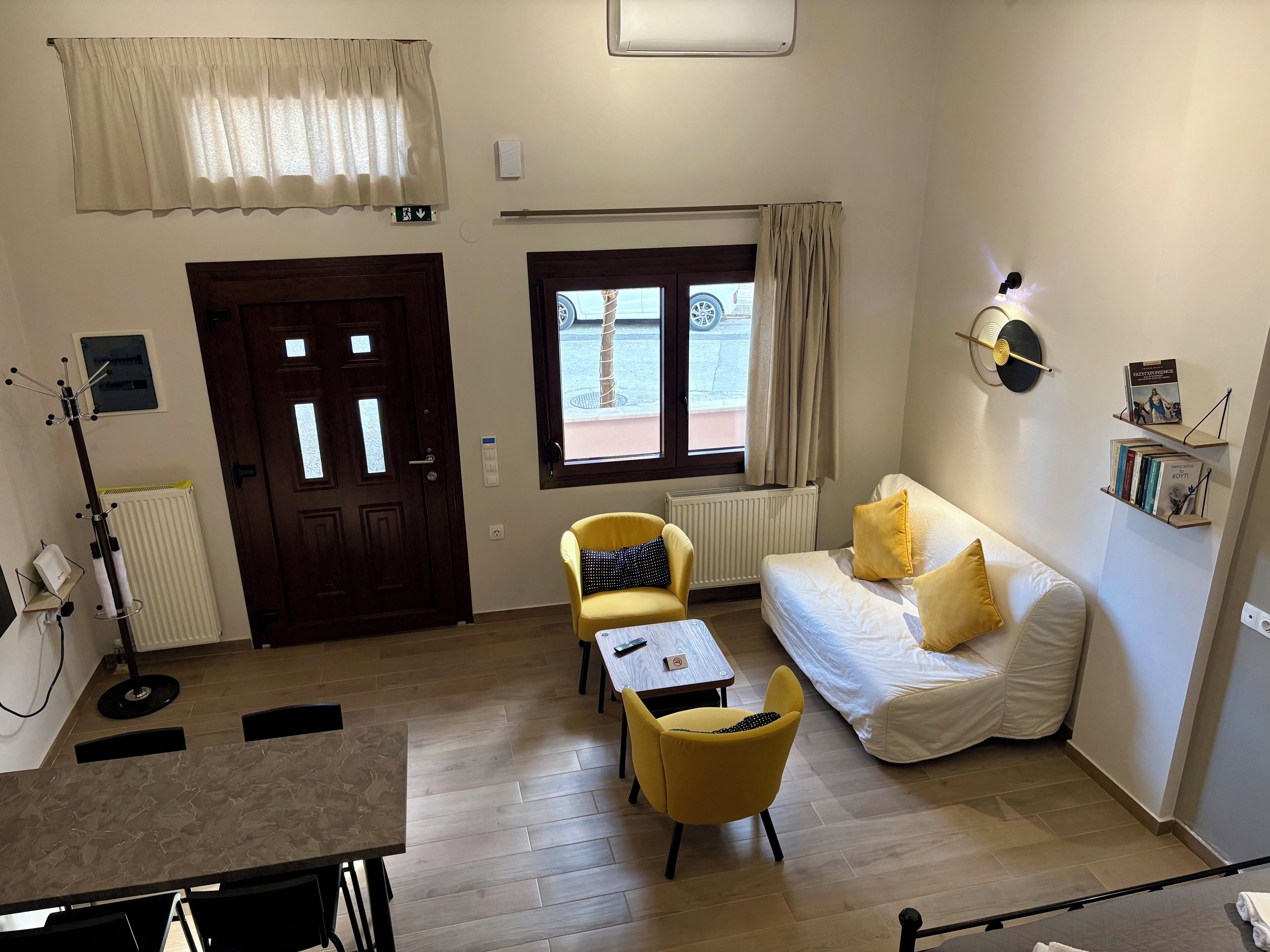 Une chambre d’hôtel avec un plancher en bois, un canapé blanc, un fauteuil jaune, une petite table, un radiateur, une porte et une fenêtre donnant sur l’extérieur.