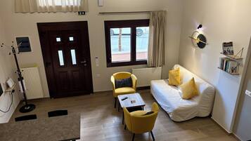 Une chambre d’hôtel avec un plancher en bois, un canapé blanc, un fauteuil jaune, une petite table, un radiateur, une porte et une fenêtre donnant sur l’extérieur.