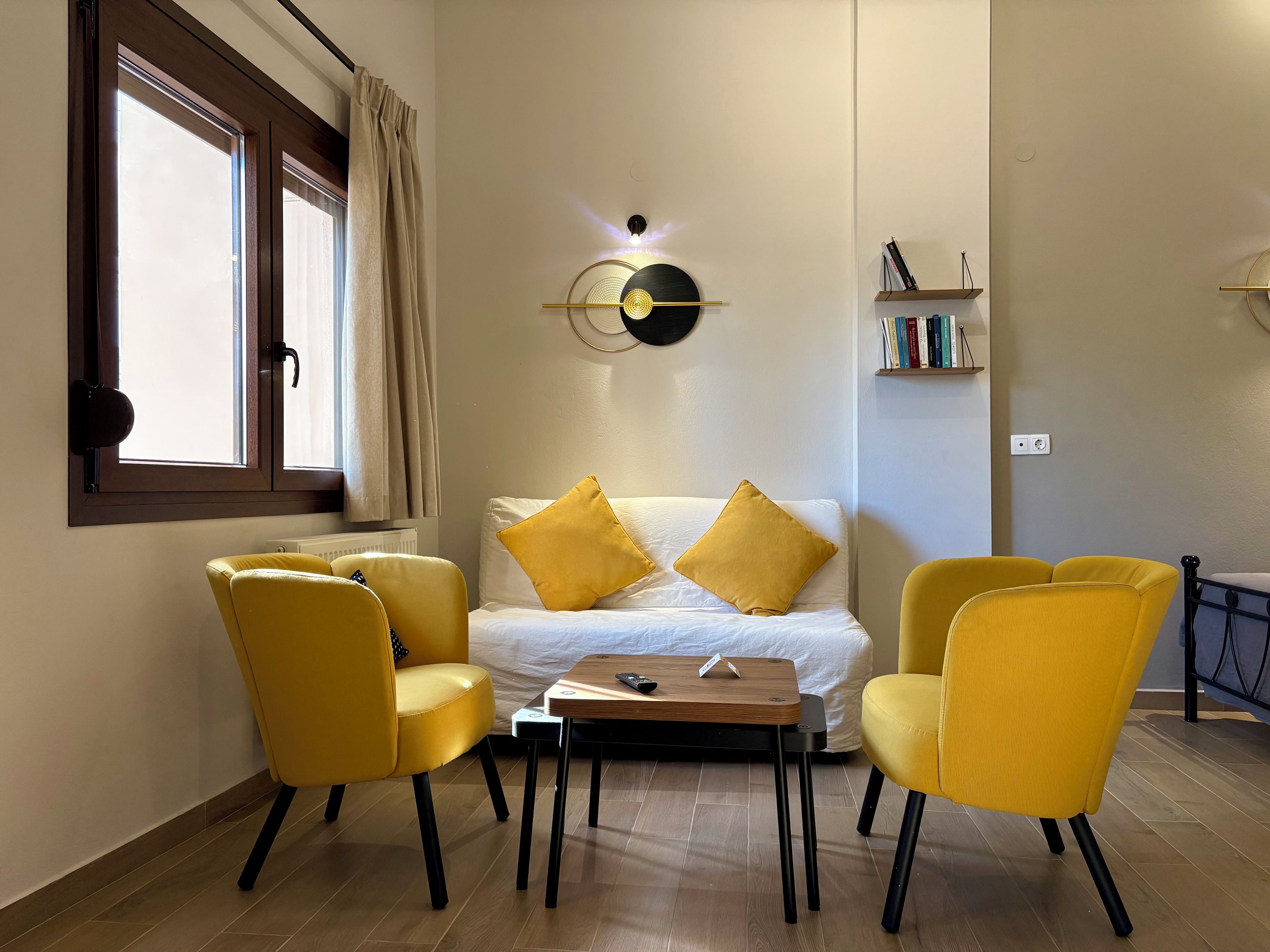 Une pièce comprenant un lit, deux chaises jaunes, une petite table, une horloge murale et une bibliothèque.