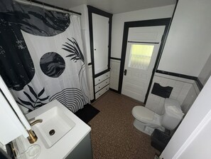 A bathroom with a white sink, a toilet, a shower curtain with a leaf pattern, and a tiled floor. - Stay Awhile • King Bed • Pet Friendly • 2BR (Elyria)