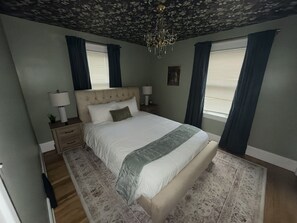 A bedroom with a large bed, a nightstand, a lamp, a chandelier, and a window with curtains. - Stay Awhile • King Bed • Pet Friendly • 2BR (Elyria)
