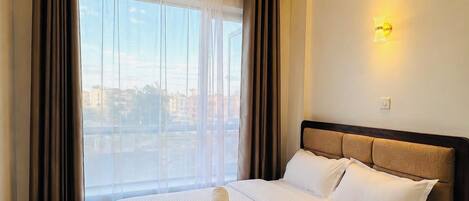 A hotel room with a large bed, a view of the city through the window, and a wall-mounted light fixture.
