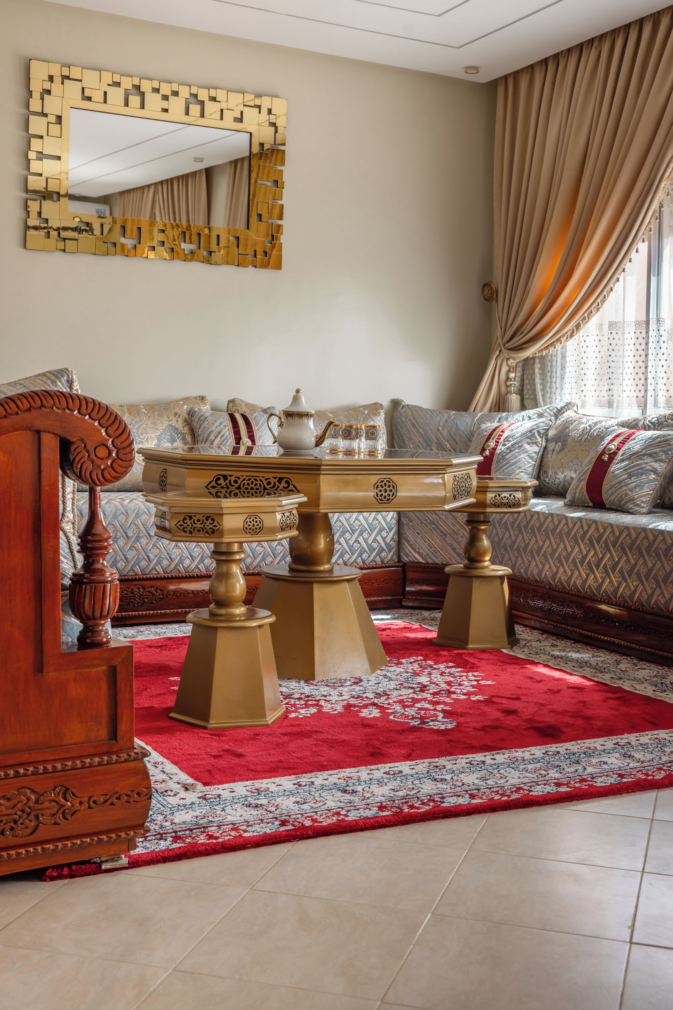 A room with a round wooden coffee table with ornate legs, a sofa with patterned cushions, and a red and white patterned rug.