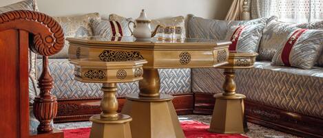 A room with a round wooden coffee table with ornate legs, a sofa with patterned cushions, and a red and white patterned rug.